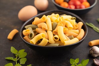 Rigatoni colored raw pasta with tomato, eggs, spices, herbs on black concrete background. Side