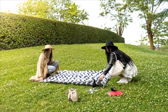Side view of a two stylish friends spreading a blanket on the lawn of a park to spend the day