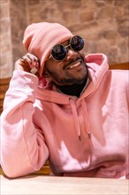 Vertical close-up portrait of a chill african man with pink urban clothes smiling sitting in a bar