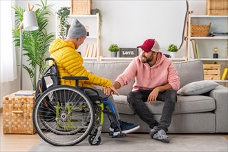 Man supporting a disabled friend in wheelchair while sitting on the sofa at home