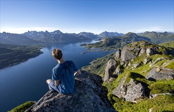 Mountaineer sitting on the summit of Dronningsvarden or Stortinden, behind mountains and fjord