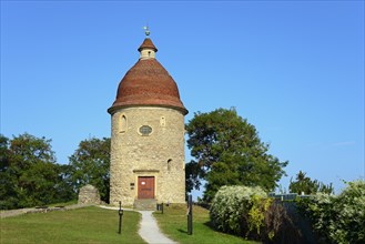 An old round tower with a red roof under a clear blue sky surrounded by green foliage, Rotunda of