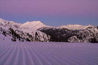 Snow-covered mountains at dusk with a purple sky and a freshly groomed ski slope in the foreground,