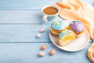 Purple and blue glazed donut and cup of coffee on blue wooden background and orange linen textile.