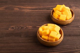 Dried and candied mango cubes in wooden bowls on brown wooden textured background. Side view, copy