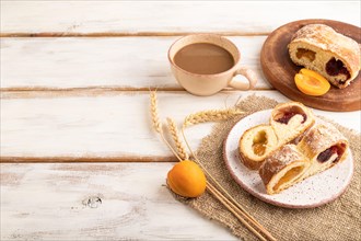 Homemade sweet bun with apricot jam and cup of coffee on white wooden background and linen textile.