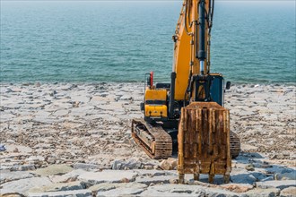 Backhoe sitting on rocky shore of man made dike with ocean in background