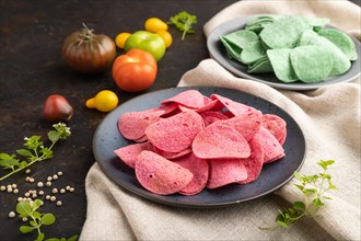Green and red potato chips with herbs and tomatoes on black concrete background and linen textile.