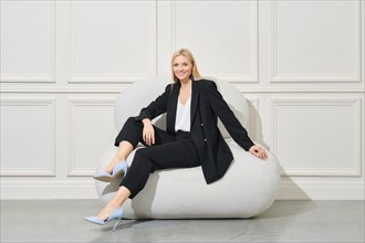 Smiling businesswoman in stylish suit relaxing on black bean bag in bright room