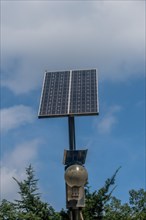 Small solar panel on top of light pole above treetops against blue cloudy sky