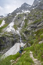 Mountaineer on a hiking trail, mountain landscape with small waterfall and rocky mountain peak,