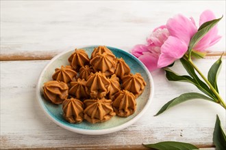 Homemade soft caramel fudge candies on blue plate on white wooden background, peony flower