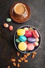 Multicolored macaroons and chocolate eggs in ceramic bowl, cup of coffee on black concrete
