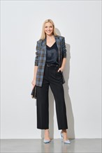 Cheerful smiling blonde woman in stylish checkered jacket and wide trousers posing by white wall