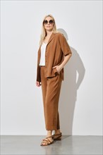 Stylish blonde woman in sunglasses posing in loose brown trousers and jacket indoors