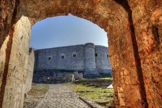 View through an old stone arch onto a sunlit fortress under a clear blue sky, Chlemoutsi, High