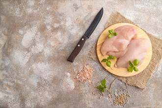 Raw chicken breast with herbs and spices on a wooden cutting board on a brown concrete background.