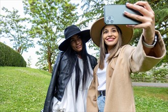 Front view of two stylish friends with hats standing and taking a selfie at the park