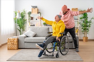 Happy disabled man in wheelchair and friend celebrating raising hands at home