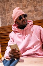 Vertical portrait of a cool african man wearing pink clothes using phone sitting in a bar