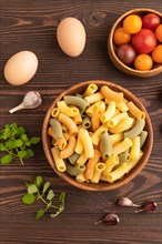 Rigatoni colored raw pasta with tomato, eggs, spices, herbs on brown wooden background. Top view,