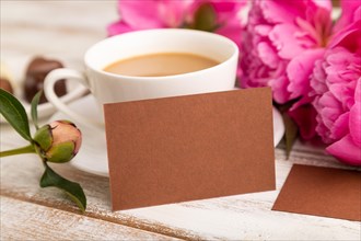 Brown business card with pink peony flowers and cup of coffee on white wooden background. side