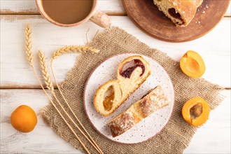 Homemade sweet bun with apricot jam and cup of coffee on white wooden background and linen textile.