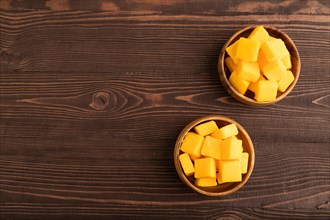 Dried and candied mango cubes in wooden bowls on brown wooden textured background. Top view, flat