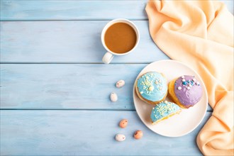 Purple and blue glazed donut and cup of coffee on blue wooden background and orange linen textile.