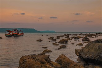 Twilight over a rocky shoreline with anchored boats and a calm sea under a soft orange sky. Koh