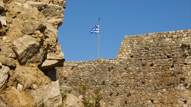 Part of an old fortress wall with a waving Greek flag and blue sky, Chlemoutsi, High Medieval