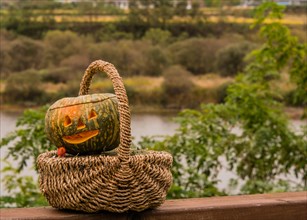 Jack-O-Lantern in wicker basket sitting on wooden railing with river and trees blurred out in