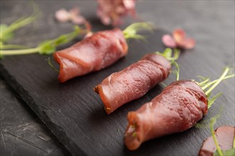 Slices of smoked salted meat with green pea microgreen on black concrete background. Side view,