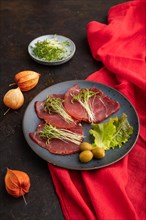 Slices of smoked salted meat with cilantro microgreen on black concrete background and red textile.