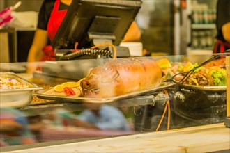 A whole roasted pig on a tray at a market stall with other dishes in the background