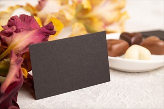 Black business card with chocolate candies and iris flowers on gray concrete background. side view,