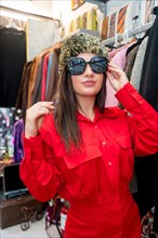 Vertical portrait of a stylish woman trying on a hat and sunglasses in a retro clothes store