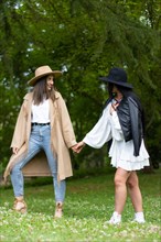 Front view of a stylish lesbian couple in hat holding hands and walking in the park while looking