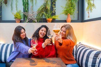 Horizontal photo of three female friends toasting with white wine sitting in a modern colorful