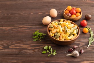 Rigatoni colored raw pasta with tomato, eggs, spices, herbs on brown wooden background. Side view,