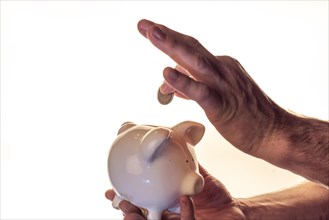 Hand of a man holding a white piggy bank and putting a coin into it, white background, studio shot,