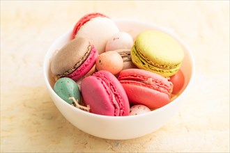 Multicolored macaroons and chocolate eggs in ceramic bowl on beige concrete background. side view,