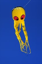A yellow kite in the shape of an octopus with many arms and red eyes in front of a blue sky