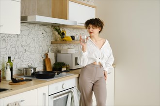 Young woman wearing a white shirt tied at the waist and beige trousers standing in the kitchen with