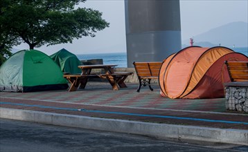 Tents set up on walkway next to picnic table and benches with the ocean and a bridge support column