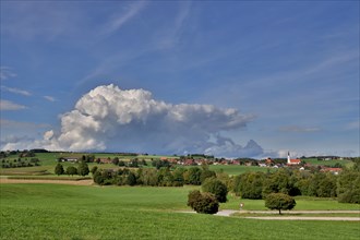 Threatening thundercloud over a sunny, hilly landscape with a village in the foothills of the Alps,