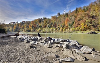 An angler sits on large stones by a river in autumn, with a castle perched on a wooded hill in the