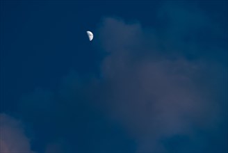 The waxing crescent moon stands bright in a deep blue evening sky with cumulus clouds illuminated