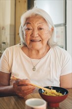 Elderly Asian woman eating rice with chopsticks looks and smiles at camera