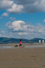 Red and black lighthouse on tidal flats during low tide with mountains and cloudy blue sky in